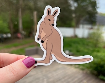 May include: A sticker featuring a cartoon kangaroo in shades of brown and black. The kangaroo is depicted in a side profile, with its tail extended. The sticker has a white border and is held against a blurred outdoor background.