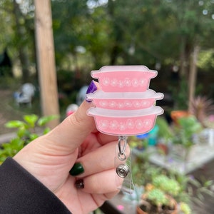 May include: A pink and white floral patterned badge holder with a retractable clip. The badge holder is shaped like a stack of three bowls.