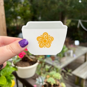 May include: White ceramic sticker with a yellow flower design. The sticker is shaped like a bowl.