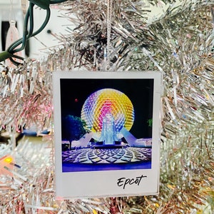 May include: A clear, square ornament featuring a photograph of the Epcot ball at night. The word "Epcot" is written in black script at the bottom. The ornament hangs from a silver tinsel Christmas tree.