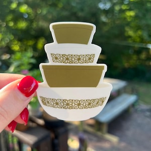 May include: A sticker featuring a stack of three decorative bowls in white and olive green. The bowls have a retro design with a floral pattern around the rims. The sticker is held up by a hand with red nail polish.