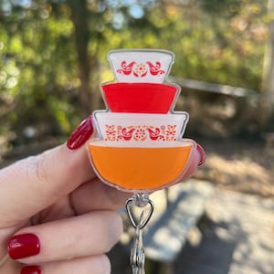 May include: A clear plastic badge holder with a stack of three colorful bowls. The bowls are orange, red, and white with a floral pattern and birds. The badge holder has a metal clip.