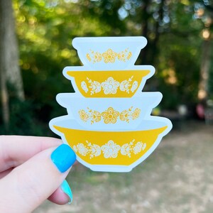 May include: A sticker featuring a stack of four yellow and white mixing bowls with a floral design. The bowls are in a nested arrangement, with the largest at the bottom and the smallest at the top. The sticker has a white border.