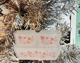 May include: A white ceramic Christmas ornament shaped like a vintage casserole dish with a pink floral design. The ornament features a repeating pattern of pink flowers and green leaves.
