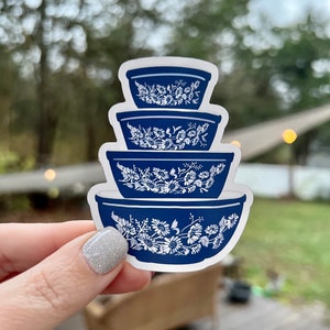 May include: A sticker featuring a stack of four blue mixing bowls with white floral designs. The bowls are nested, with the largest at the bottom and the smallest at the top. The sticker is held against a blurred outdoor background.