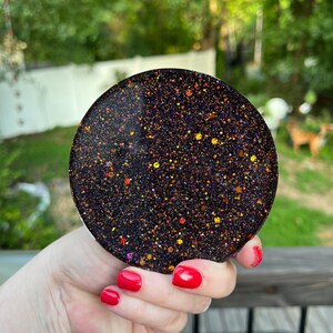 May include: A round black coaster with gold and orange glitter embedded in the resin. The coaster is being held by a hand with red nail polish.