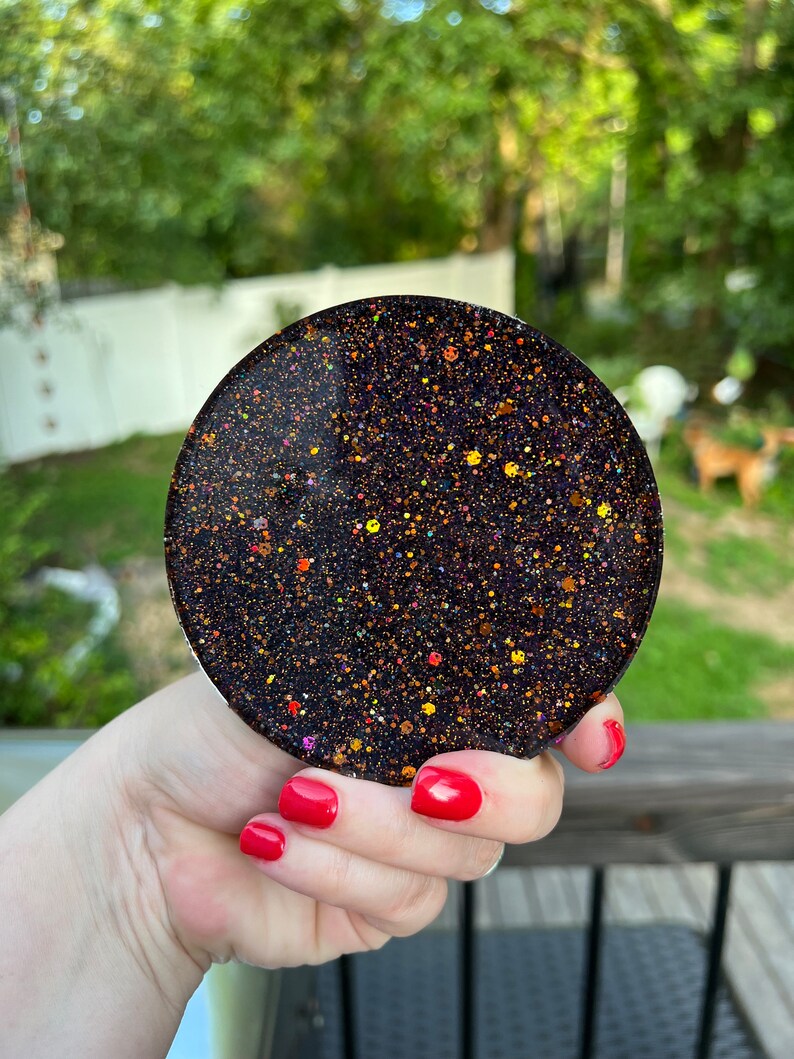 May include: A round black coaster with gold and orange glitter embedded in the resin. The coaster is being held by a hand with red nail polish.