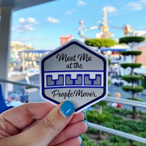 May include: A silver hexagon sticker with blue accents. The sticker says "Meet Me at the People Mover" with a stylized graphic of a train.