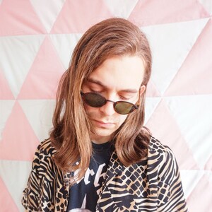 May include: A person wearing a brown and black patterned shirt with a black t-shirt underneath. The t-shirt has a graphic print of a skull with horns. The person is wearing round, brown sunglasses.