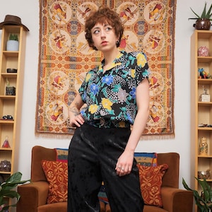 May include: A woman wearing a black and white floral print short-sleeved shirt with a black satin pants. She is standing in front of a tapestry and a bookshelf.