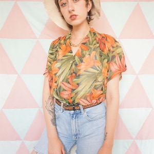 May include: A woman wearing a sheer, orange and green floral print button-up shirt with a brown belt, light blue denim shorts, and a straw hat. She has a tattoo on her left leg.