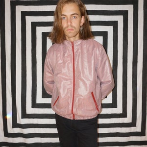 May include: A person wearing a pink striped jacket with a red zipper and black pants. The person is standing in front of a black and white striped background.