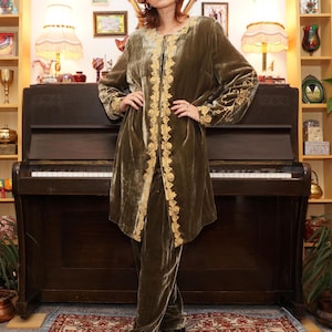 Op de afbeelding: Een lange, olijfgroene fluwelen jas met gouden borduurwerk. De jas heeft een shawlkraag en een voorsluiting met een knoop. De jas wordt gedragen over een olijfgroene broek.