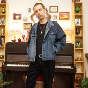 May include: A man wearing a blue denim jacket, a black t-shirt, black pants with white stripes, and a silver necklace with a pendant. He is standing in front of a piano with a guitar leaning against a shelf in the background.
