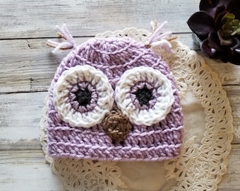May include: A purple crocheted owl hat with white and black eyes and a brown nose. The hat is sitting on a white crocheted doily.