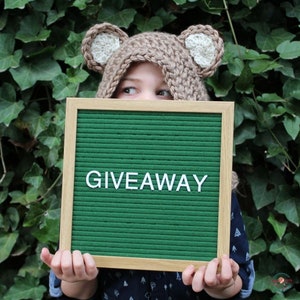 May include: A child wearing a brown crocheted bear hat with white ears holds a green letter board with the word "GIVEAWAY" spelled out in white letters.