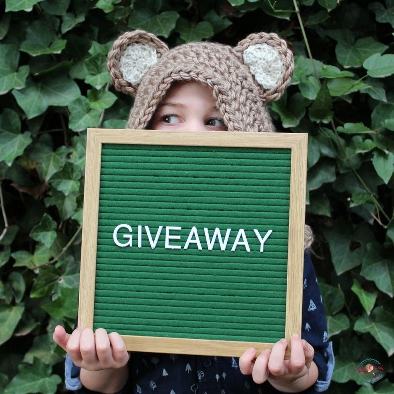 May include: A child wearing a brown crocheted bear hat with white ears holds a green letter board with the word "GIVEAWAY" spelled out in white letters.