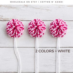 May include: Three round yarn pom-poms featuring a mix of shocking pink, baby pink, and white fibres. Each pom-pom has long white yarn tails extending downwards. Text on the image reads 2 Colors + White POM-POMS and SHOWN: SHOCKING PINK & BABY PINK.