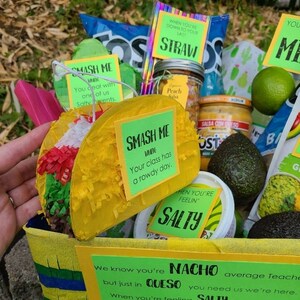 May include: A yellow and blue basket filled with a variety of snacks and a taco pi&ntilde;ata. The basket is decorated with a sign that reads "NACHO average Teacher, but just in QUESO you need us we're here. When you're feeling SALTY and need to TACO 'bout something, just call. Find your happy MIX and remember CACTUS makes perfect."