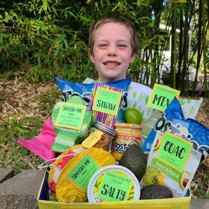 May include: A yellow and green gift basket filled with snacks and a variety of items with labels that say "Smash Me", "When You're Feeling Salty", "Your Happy Mix", "Straw", and "Guac". The basket is decorated with yellow, blue, and green streamers. The text on the basket reads "We know you're Nacho average Teacher, but just in Queso you need us, we're here. When you're feeling Salty and need to Taco 'bout something just call. Find your happy Mix and remember Cactus makes me happy."