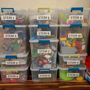 以下が含まれることがあります： STEM 1～12のラベルが付いた透明なプラスチック製の収納箱で、おもちゃが詰まっています。箱には青と緑のハンドルが付いており、木製の床に積み重ねられています。上部には「Toy Rotation Printables from A Cotton Kandi Life」と書かれています。