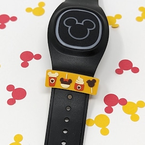 May include: A black MagicBand with a Mickey Mouse icon on the front. The band has a yellow band with a Mickey Mouse head, a Mickey Mouse pretzel, a Mickey Mouse ice cream cone, and a Mickey Mouse coffee cup on it. The band is surrounded by red and yellow Mickey Mouse head confetti.