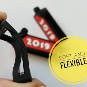 K&ouml;nnte beinhalten: Zwei schwarze und rote Gummianh&auml;nger mit dem Jahr 2019 darauf gedruckt. Die Anh&auml;nger sind flexibel und biegsam. Der Text "SOFT AND FLEXIBLE" ist auf einem gelben Kreis gedruckt.