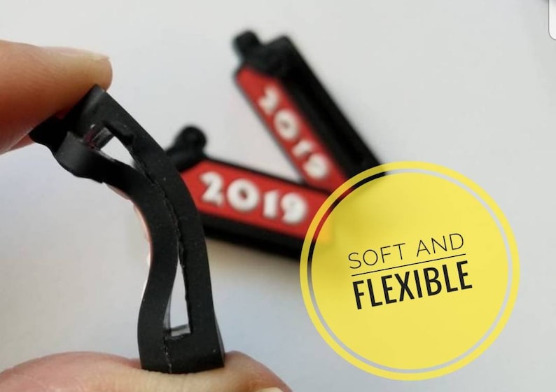 K&ouml;nnte beinhalten: Zwei schwarze und rote Gummianh&auml;nger mit dem Jahr 2019 darauf gedruckt. Die Anh&auml;nger sind flexibel und biegsam. Der Text "SOFT AND FLEXIBLE" ist auf einem gelben Kreis gedruckt.