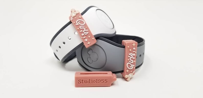 K&ouml;nnte beinhalten: Zwei rosa und wei&szlig;e Disney MagicBands mit einem rosa Anh&auml;nger, auf dem jeweils "Girls Trip" steht. Der Anh&auml;nger ist mit einem goldenen Mickey-Maus-Kopf am Band befestigt.