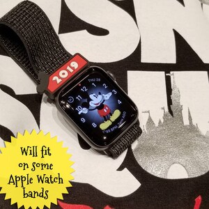 K&ouml;nnte beinhalten: Eine schwarze Apple Watch mit einem Mickey-Maus-Gesicht auf dem Bildschirm. Die Uhr ist an einem schwarzen Armband und hat einen roten Mickey-Maus-Kopf-Armband-Anh&auml;nger. Die Uhr liegt auf einem schwarzen Shirt mit dem Wort "Disney" in Rot gedruckt. Der Text "2019" ist auf dem Armband-Anh&auml;nger gedruckt. Der Text "Will fit on some Apple Watch bands" ist auf einem gelben Stern-Aufkleber gedruckt.