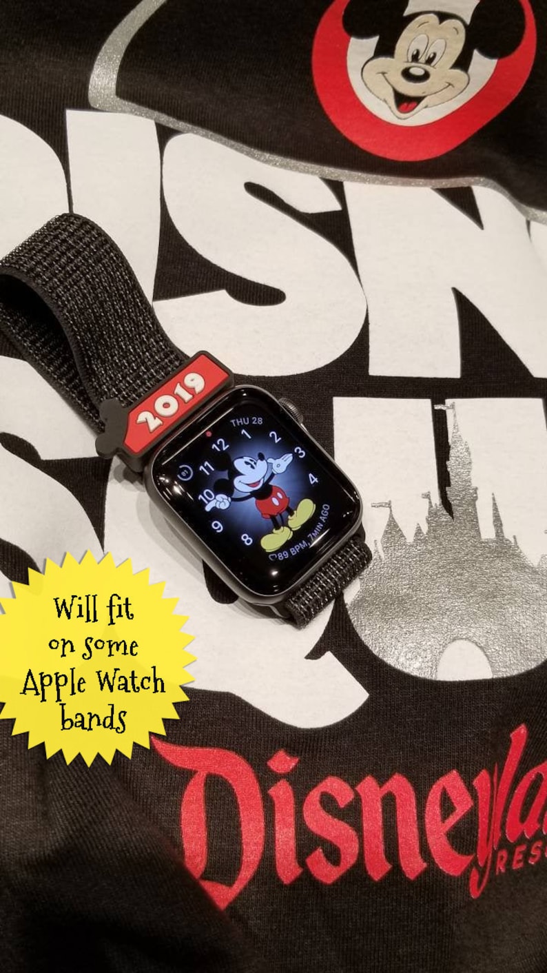 K&ouml;nnte beinhalten: Eine schwarze Apple Watch mit einem Mickey-Maus-Gesicht auf dem Bildschirm. Die Uhr ist an einem schwarzen Armband und hat einen roten Mickey-Maus-Kopf-Armband-Anh&auml;nger. Die Uhr liegt auf einem schwarzen Shirt mit dem Wort "Disney" in Rot gedruckt. Der Text "2019" ist auf dem Armband-Anh&auml;nger gedruckt. Der Text "Will fit on some Apple Watch bands" ist auf einem gelben Stern-Aufkleber gedruckt.