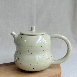Op de afbeelding: Een lichtcrèmekleurige keramische theepot met een gespikkeld bruin patroon over het lichaam, het deksel en het handvat. De theepot heeft een ronde, bolvormige vorm met een korte tuit en een bijpassend deksel met een kleine knop.