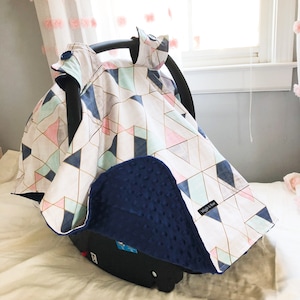 May include: A baby car seat cover with a geometric pattern in pink, blue, and gold on a white background. The cover has a navy blue, textured underside and is secured with a large blue button.