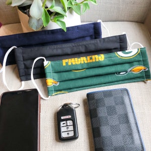 May include: Three face masks in navy blue, black, and green with white ear loops. The green mask has a yellow and white design. A black smartphone, car key fob, and a black and gray wallet are also visible.