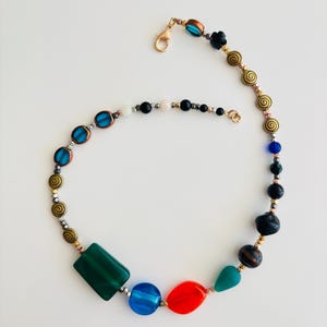 May include: A colorful beaded necklace with a variety of shapes and colors. The necklace features a mix of green, blue, red, and gold beads, with a gold clasp. The design includes geometric and spiral-shaped beads.
