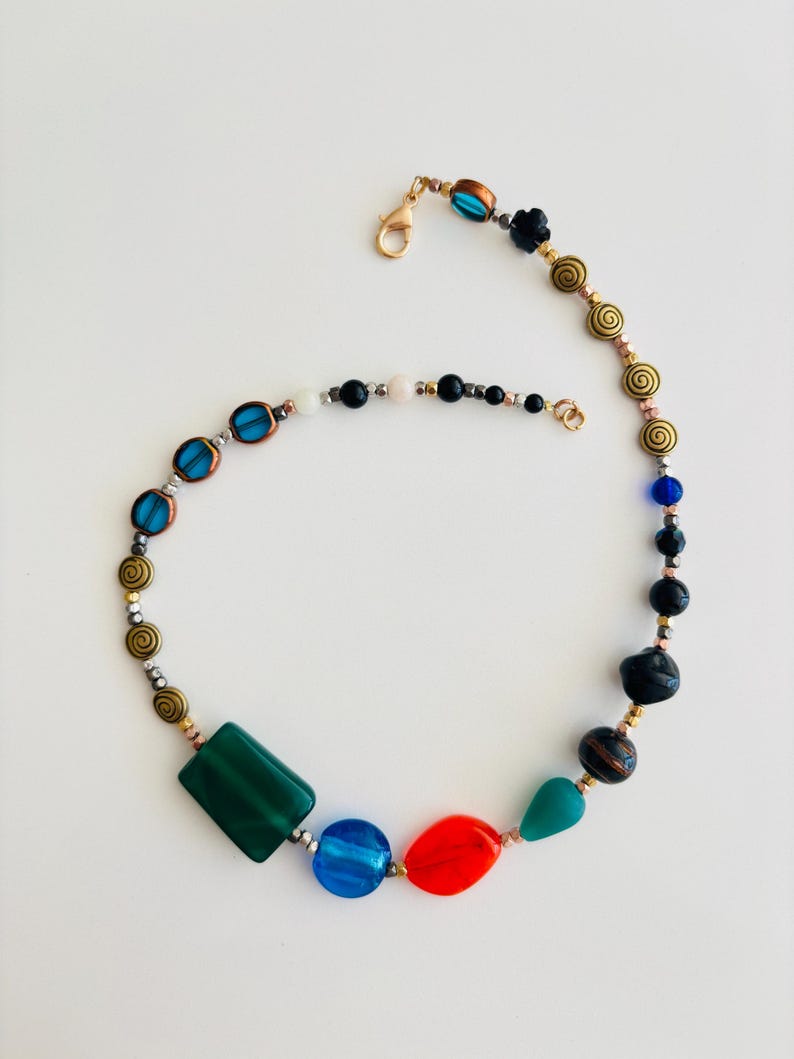 May include: A colorful beaded necklace with a variety of shapes and colors. The necklace features a mix of green, blue, red, and gold beads, with a gold clasp. The design includes geometric and spiral-shaped beads.