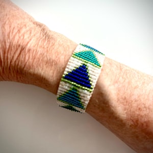 May include: A beaded bracelet with a geometric pattern of blue, green, and white triangles. The bracelet is worn on a wrist and has a woven appearance. The design features a repeating pattern of triangles in shades of blue and green, set against a white background.