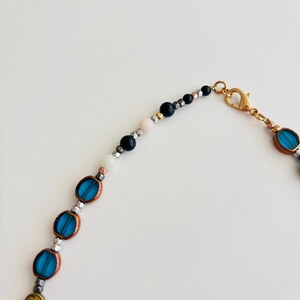 May include: A beaded necklace featuring oval turquoise beads, metallic silver beads, and gold spiral beads. The necklace has a gold clasp and a mix of black, white, and pink beads near the clasp. The beads are strung on a thin, flexible cord.