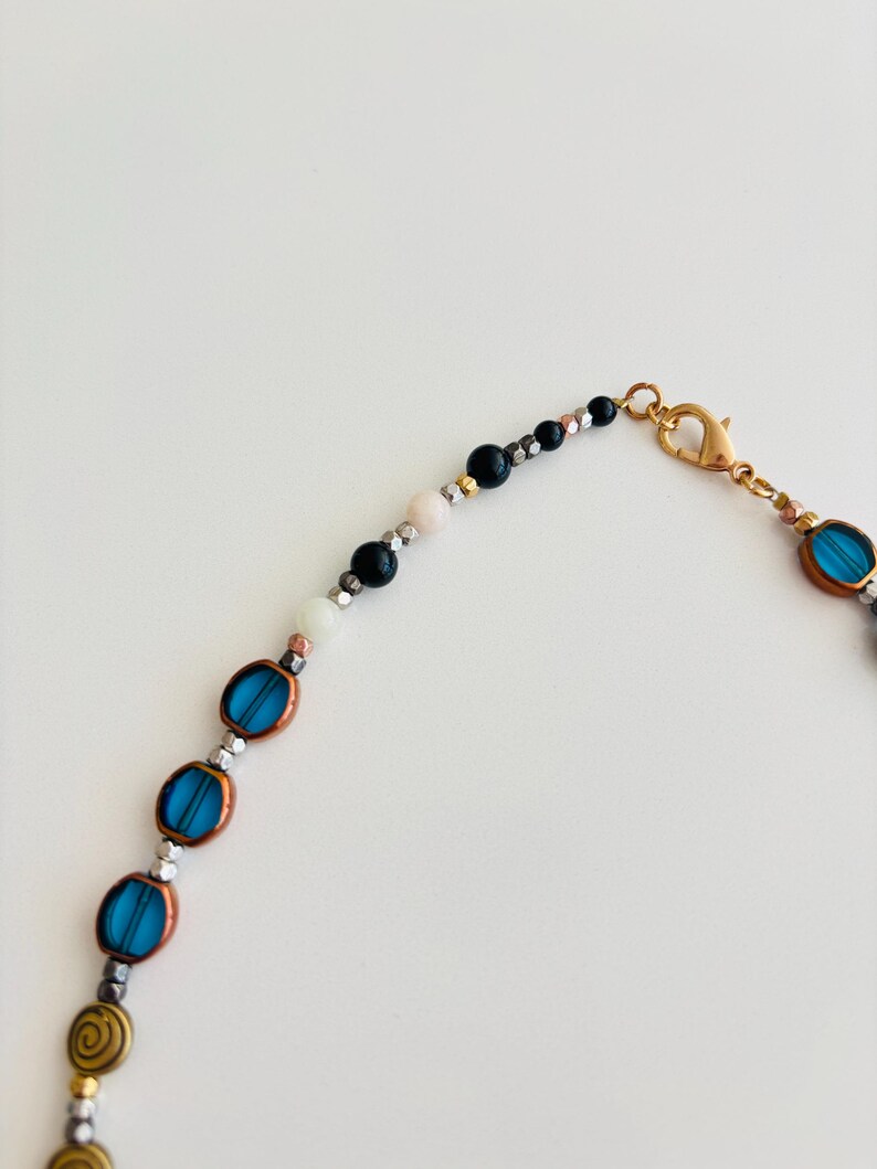 May include: A beaded necklace featuring oval turquoise beads, metallic silver beads, and gold spiral beads. The necklace has a gold clasp and a mix of black, white, and pink beads near the clasp. The beads are strung on a thin, flexible cord.
