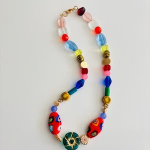 May include: A colorful beaded necklace with a variety of shapes and sizes. The necklace features red, blue, green, yellow, and gold beads. The focal point includes red oval beads with floral designs and a green flower-shaped bead.