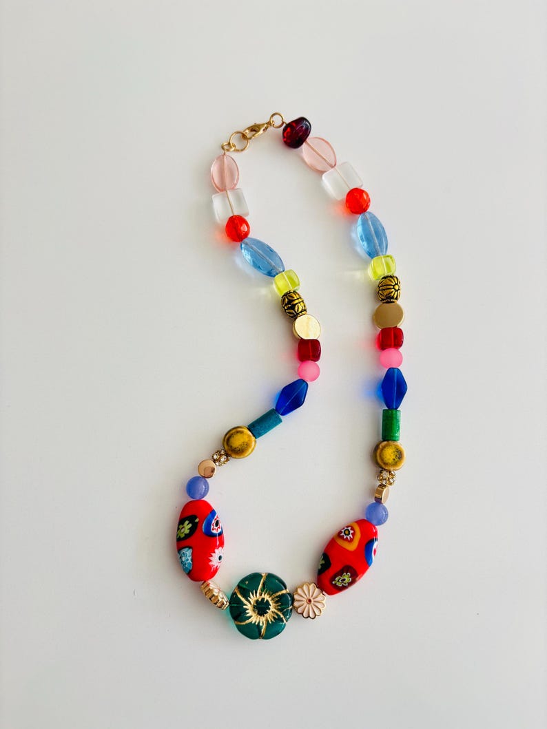 May include: A colorful beaded necklace with a variety of shapes and sizes. The necklace features red, blue, green, yellow, and gold beads. The focal point includes red oval beads with floral designs and a green flower-shaped bead.
