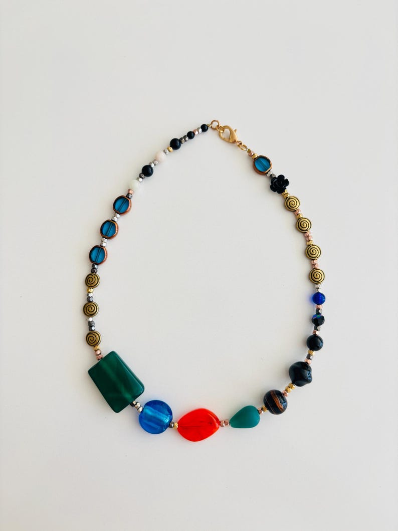 May include: A colorful beaded necklace featuring a variety of shapes and colors. The necklace includes green, blue, red, and black beads, as well as gold-toned spiral and round beads. The necklace is on a white background.