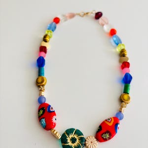May include: A colorful beaded necklace with a variety of shapes and sizes. The necklace features red, blue, green, yellow, and gold beads. Two large red oval beads with floral designs are prominent. A green flower-shaped bead and a small pink flower accent the design.