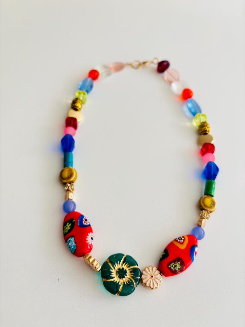 May include: A colorful beaded necklace with a variety of shapes and sizes. The necklace features red, blue, green, yellow, and gold beads. Two large red oval beads with floral designs are prominent. A green flower-shaped bead and a small pink flower accent the design.