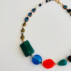 May include: A colorful necklace featuring a variety of beads in shades of green, blue, red, and gold. The necklace includes rectangular, round, and teardrop-shaped beads, as well as spiral and small round beads. The clasp is gold-toned.