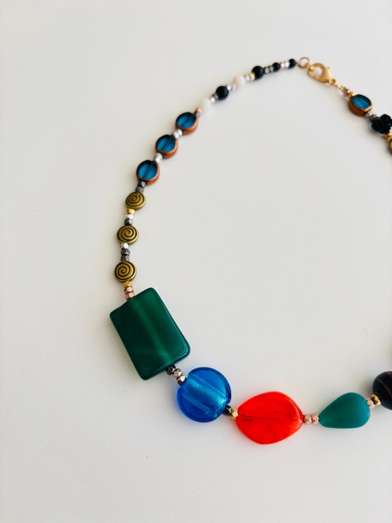 May include: A colorful necklace featuring a variety of beads in shades of green, blue, red, and gold. The necklace includes rectangular, round, and teardrop-shaped beads, as well as spiral and small round beads. The clasp is gold-toned.