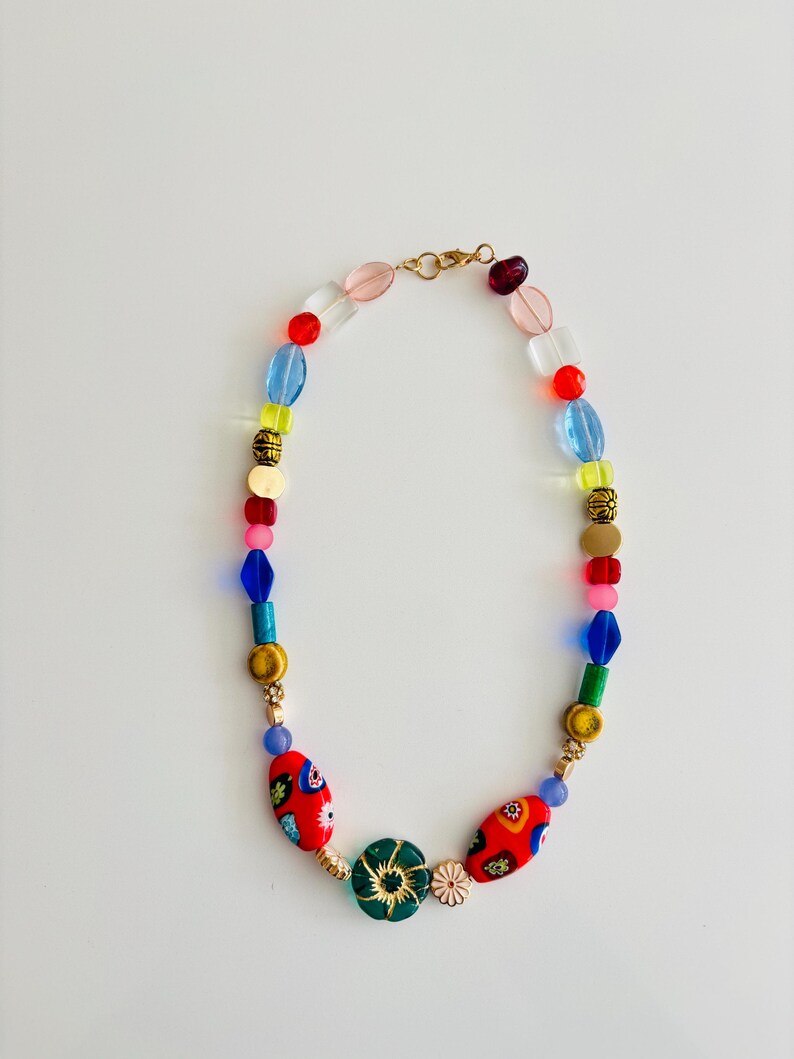 May include: A colorful beaded necklace with a gold clasp. The necklace features a variety of beads in red, blue, green, yellow, and pink. Some beads have floral patterns. The necklace is on a white background.