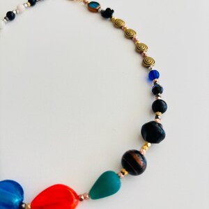 May include: A colorful necklace featuring a variety of beads in blue, red, green, black, and gold. The necklace has a gold-tone clasp and includes spiral and oval-shaped beads.