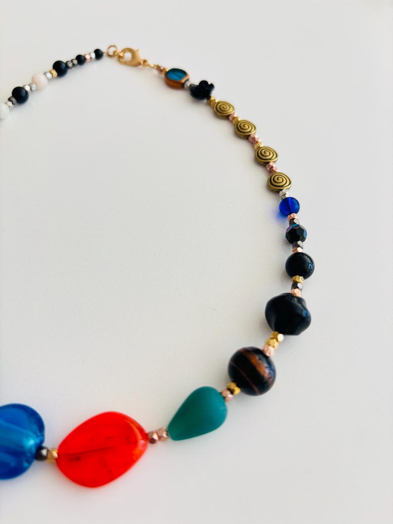 May include: A colorful necklace featuring a variety of beads in blue, red, green, black, and gold. The necklace has a gold-tone clasp and includes spiral and oval-shaped beads.