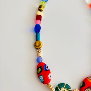 May include: A colorful beaded necklace with a variety of shapes and sizes. The necklace features red, blue, green, and gold beads, with a gold clasp. The focal point includes red oval beads with floral designs.